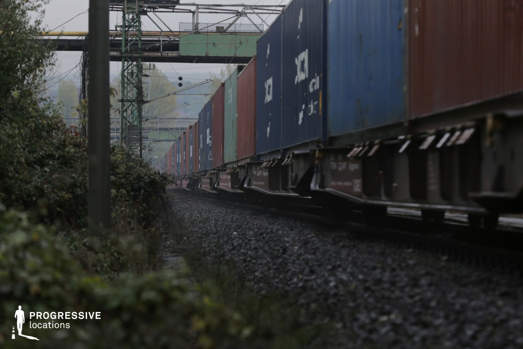 This dynamic railway scene captures a long freight train carrying stacked shipping containers as it moves through an industrial corridor. The steel tracks, embedded in dark gravel, curve gently into the distance, adding depth and motion. Overhead, rusted metal bridges and electrical lines crisscross the frame, creating a layered, mechanical aesthetic. The surrounding vegetation, creeping along the edges of the railway, contrasts with the industrial setting, hinting at nature reclaiming space. This location is ideal for filming gritty urban dramas, industrial thrillers, or transportation-themed sequences, offering a visually rich environment with a raw, atmospheric quality.
