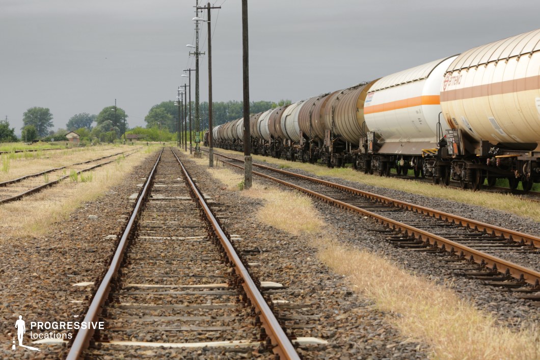 This expansive railway setting features a long freight train carrying cylindrical tanker cars, stretching into the distance along parallel tracks. The gravel-lined rails, flanked by patches of dry grass, create a rugged, industrial aesthetic. Wooden utility poles with overhead lighting run alongside the tracks, reinforcing the sense of vast, open infrastructure. Small rural buildings and scattered trees in the background contrast with the heavy machinery, adding a remote and isolated atmosphere. This location is well-suited for filming industrial, western-inspired, or neo-noir scenes, capturing themes of solitude, long-distance travel, and logistical movement in a raw, cinematic landscape.
