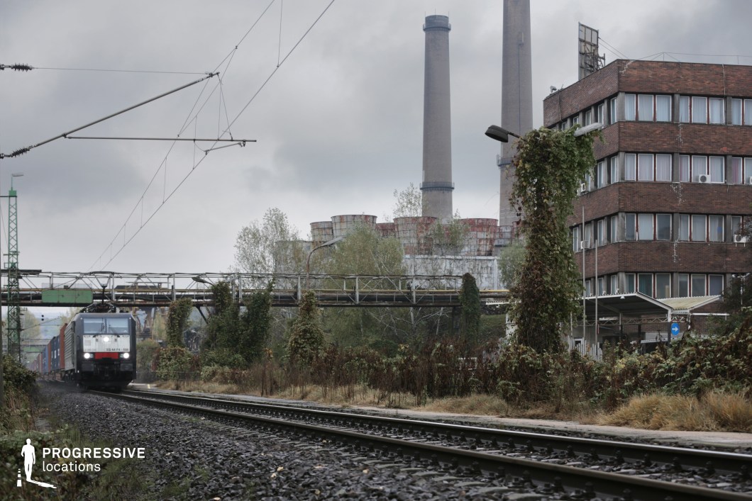 This railway scene is set against an imposing industrial landscape, featuring towering smokestacks, rusted storage tanks, and a weathered brick office building. The train, hauling freight containers, moves along steel tracks embedded in dark gravel, reinforcing the heavy, utilitarian feel of the location. Overhead power lines and a vine-covered pedestrian bridge add layers of depth and structure, contrasting the rigid man-made elements with encroaching nature. The blend of industrial decay and active infrastructure makes this setting ideal for filming dystopian, neo-noir, or gritty urban dramas, offering a raw and atmospheric visual aesthetic.