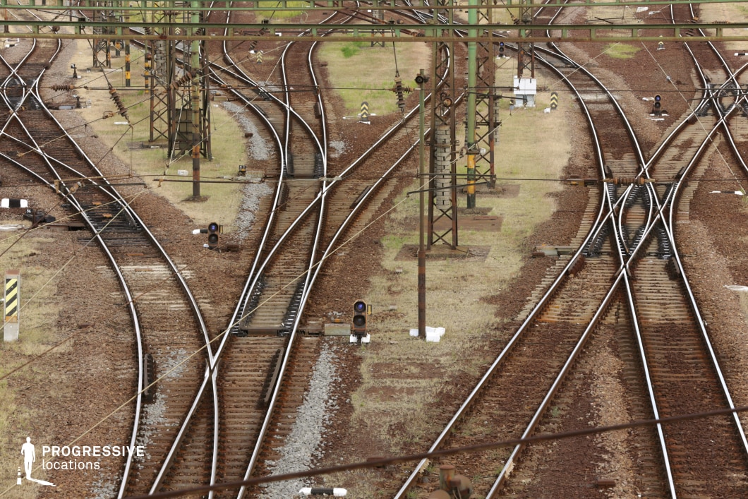 This intricate railway junction presents a visually striking network of converging and diverging tracks, creating a labyrinthine pattern of steel rails embedded in reddish-brown gravel. Overhead electrical wires crisscross the scene, supported by tall, weathered metal poles, adding an industrial aesthetic to the structured chaos below. Signal lights and mechanical track switches are scattered throughout, reinforcing the sense of control within the complexity. The muted tones of the landscape contrast with the sharp geometry of the rails, making this an ideal filming location for themes of decision-making, tension, or movement. Perfect for thrillers, neo-noir, and urban dramas, this setting offers dynamic compositions and strong visual storytelling elements.