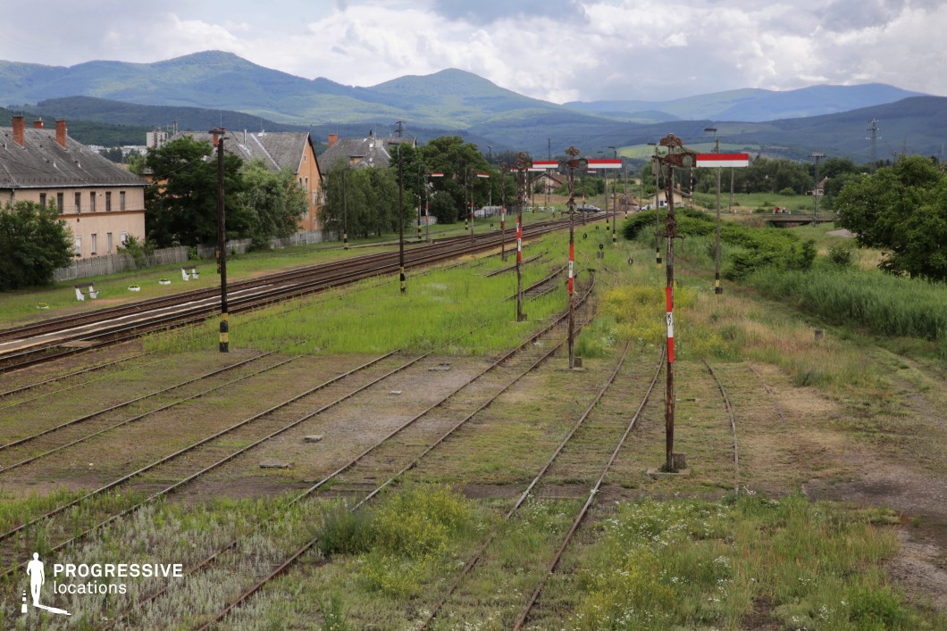 This expansive railway junction, set in a rural landscape, features multiple sets of tracks merging and diverging amidst overgrown grass and wild vegetation. A series of old semaphore signals with faded red and white markings stand along the tracks, adding a vintage and somewhat abandoned aesthetic to the scene. A row of modest buildings with pitched roofs lines the left side, reinforcing the quiet, small-town character of the location. In the background, rolling green hills and mountains create a stunning natural contrast against the man-made infrastructure. This location is ideal for filming period dramas, historical narratives, or contemplative road-movie sequences, offering a blend of nostalgia, isolation, and scenic beauty.