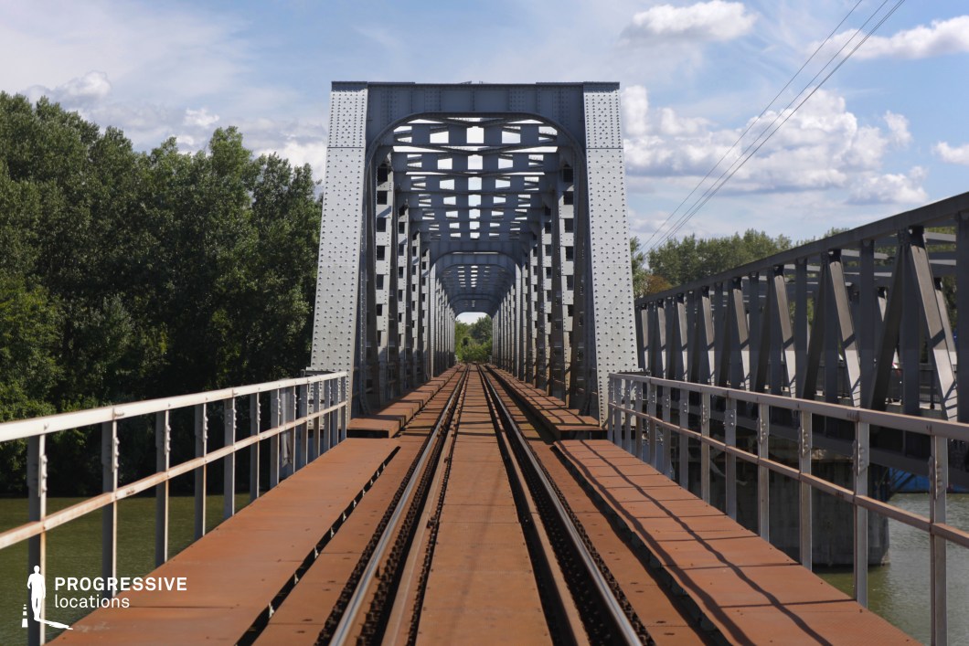 This imposing steel truss railway bridge extends over a river, its repeating geometric framework creating a striking tunnel-like perspective. The riveted metal structure, painted in a muted industrial gray, contrasts with the rust-colored tracks and the natural greenery lining the riverbanks. Strong vertical and horizontal lines guide the eye toward the distant vanishing point, emphasizing depth and symmetry. The solid railings and sturdy construction evoke a sense of durability and engineering precision. This location is ideal for filming action sequences, dramatic travel shots, or symbolic journey scenes, making it a perfect backdrop for thrillers, road movies, or historical dramas.