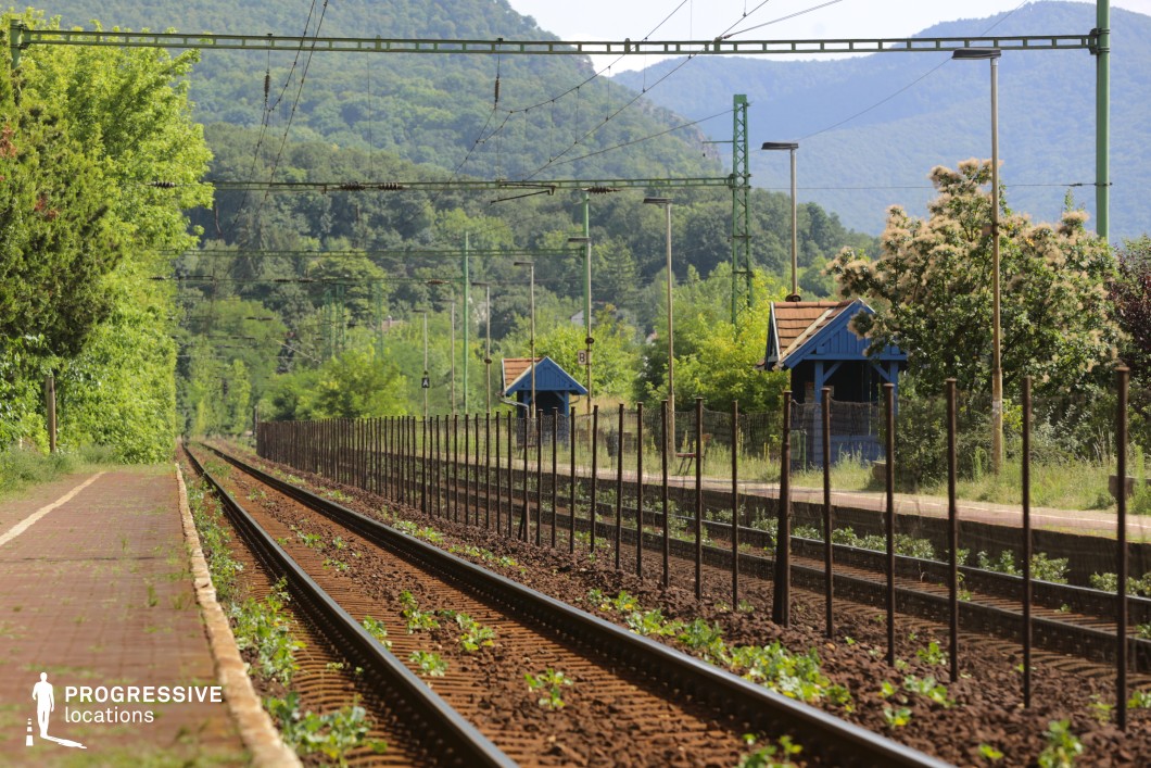 This quiet railway station is nestled in a lush, mountainous landscape, with tracks running parallel to a rustic platform. Small blue wooden shelters with sloped roofs add a quaint, old-fashioned charm, while overhead electrical lines and metal poles introduce an industrial contrast. A simple wire fence runs alongside the tracks, subtly dividing the railway from the overgrown vegetation creeping through the gravel. In the background, densely forested hills create a serene and immersive natural setting. This location is ideal for filming contemplative travel sequences, rural dramas, or nostalgic period films, offering a visually rich mix of nature and transportation infrastructure.