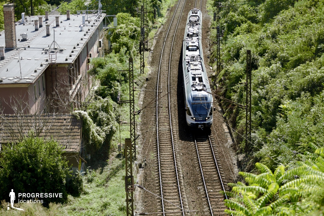 This aerial view captures a modern train traveling along a set of well-maintained railway tracks, flanked by dense vegetation and an aging residential building. Overhead power lines and steel poles frame the railway, adding an industrial contrast to the surrounding greenery. The adjacent building, with its flat rooftop and weathered facade, integrates into the scene, evoking a sense of urban history merging with nature. The railway corridor appears both functional and somewhat secluded, making it an ideal filming location for travel sequences, urban dramas, or psychological thrillers where themes of transition, isolation, or industrial aesthetics play a central role.