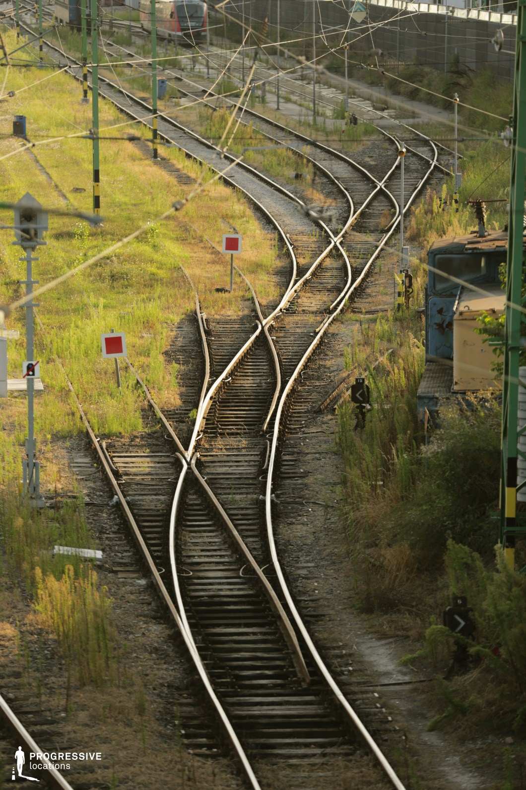 This railway junction features multiple sets of diverging and converging tracks, creating an intricate pattern of steel rails and wooden sleepers. The aged railway infrastructure, with overgrown vegetation creeping between the tracks, suggests a less frequently used or semi-abandoned section of the network. Overhead electrical wires crisscross the scene, adding a complex web of industrial elements. Red and white railway signs punctuate the landscape, marking critical switching points. The composition is framed by metal poles and a weathered train car on the right, reinforcing the raw, industrial atmosphere. This location is ideal for filming dramatic railway sequences, industrial settings, or scenes emphasizing transition and choice, making it suitable for thriller, neo-noir, or historical drama genres.