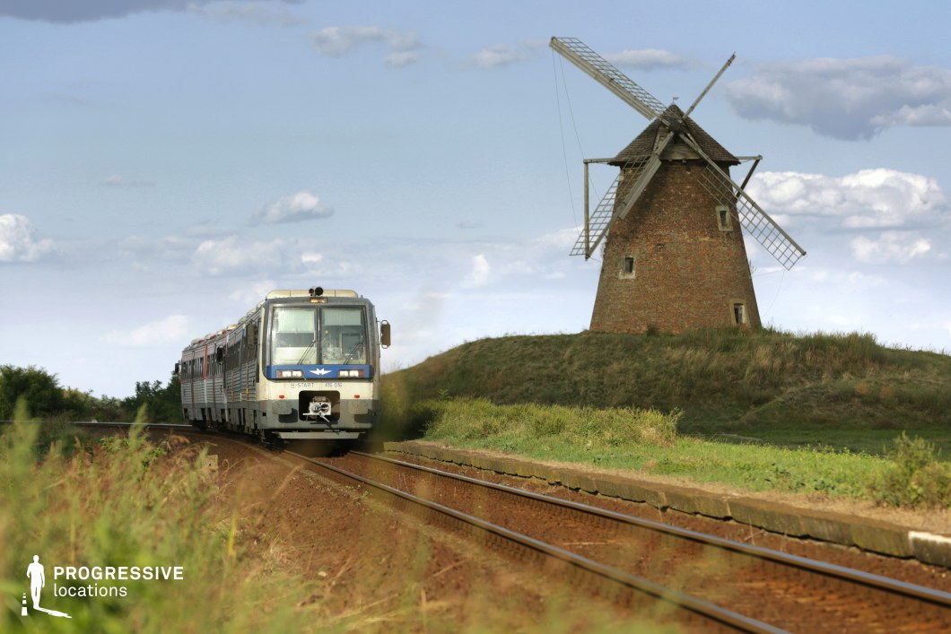 This scenic railway setting features a train moving along curved tracks through a vast, open countryside. A traditional brick windmill with wooden sails stands atop a gently sloping hill, its weathered structure evoking a sense of history and timelessness. The railway tracks, set in reddish-brown gravel, cut through the grassy landscape, creating a strong visual contrast with the green vegetation. The horizon is wide and unobstructed, emphasizing the rural openness and peacefulness of the location. This spot is ideal for filming period pieces, nostalgic travel sequences, or contemplative dramas, offering a blend of rustic charm and cinematic depth.