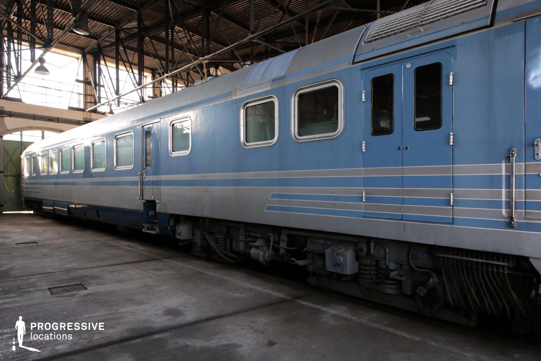 A streamlined blue train car with a metallic finish, housed inside an industrial train depot. The body features horizontal silver stripes, giving it a sleek, mid-century aesthetic. The rectangular windows are reinforced, and the heavy-duty double doors suggest a specialized or armored function. The roof has a ventilated section, possibly for air circulation or mechanical equipment. The undercarriage is equipped with robust suspension and mechanical components, indicating a well-built and durable structure. The industrial setting enhances the historical and functional character of this train, making it an ideal backdrop for period or military-themed productions.