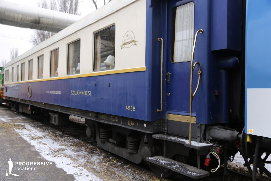 This vintage train carriage, painted in a striking blue and cream color scheme with gold accents, exudes a refined and nostalgic atmosphere. The large windows offer a glimpse into the elegantly furnished interior, while polished brass railings and fixtures enhance the luxurious aesthetic. The bold lettering and insignias on the exterior emphasize its historical significance, likely part of a heritage or luxury train service. The classic design and well-preserved condition make this an ideal filming location for period dramas, high-society travel narratives, or mystery thrillers set in an exclusive railway setting.