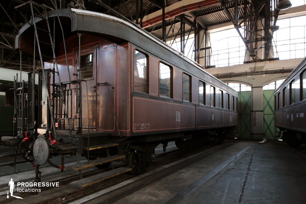This vintage railway carriage, painted in rich brown tones with fine detailing, exudes early 20th-century charm. The riveted metal exterior, tall rectangular windows, and decorative paneling highlight its classic craftsmanship. The rear platform, complete with metal railings, steps, and mechanical couplings, adds to its industrial elegance. Positioned inside a spacious train depot with exposed steel beams and large arched windows, the setting offers a dramatic and nostalgic atmosphere. This location is ideal for period dramas, noir thrillers, or historical travel narratives requiring a visually striking and historically rich train environment.