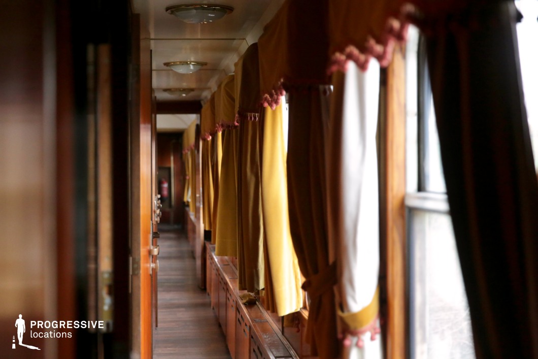 This narrow corridor inside a vintage train car features warm wooden paneling, golden drapes with decorative tassels, and large windows that allow soft, natural light to filter in. The gently swaying curtains and polished wood surfaces create an atmosphere of classic elegance, emphasizing the train's timeless charm. Overhead lighting fixtures contribute to the cozy yet sophisticated ambiance. The corridor's depth and perspective make it an ideal filming location for period dramas, mystery thrillers, or travel-themed narratives requiring a refined and nostalgic railway setting.