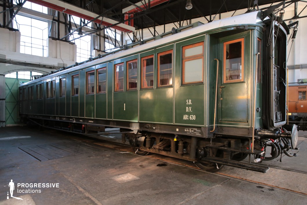 This elegant, deep green railway carriage reflects a bygone era of travel, with its polished wooden window frames and subtle gold detailing along the exterior panels. The structure is robust, featuring large riveted panels, an arched roof, and metal footboards, emphasizing its historical significance. The carriage is stationed inside an industrial depot with high ceilings and large-paned windows, allowing natural light to highlight its classic form. The soft interior curtains visible through the windows suggest a refined travel experience. This setting is ideal for period film productions, historical reenactments, or nostalgic railway-themed visuals.