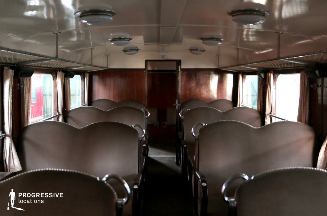 A historic mid-20th-century passenger train car with a symmetrical seating arrangement featuring curved, brown upholstered seats with chrome-finished armrests. The dark wooden paneling and cream-colored ceiling contribute to a utilitarian yet classic aesthetic. Overhead metal luggage racks line both sides of the carriage, while beige curtains with a subtle pattern frame the large windows, adding a touch of warmth. The round, evenly spaced ceiling lamps provide uniform lighting. The simple, functional design reflects a Soviet-era travel setting, making this an ideal location for period films, historical documentaries, or retro-inspired productions.