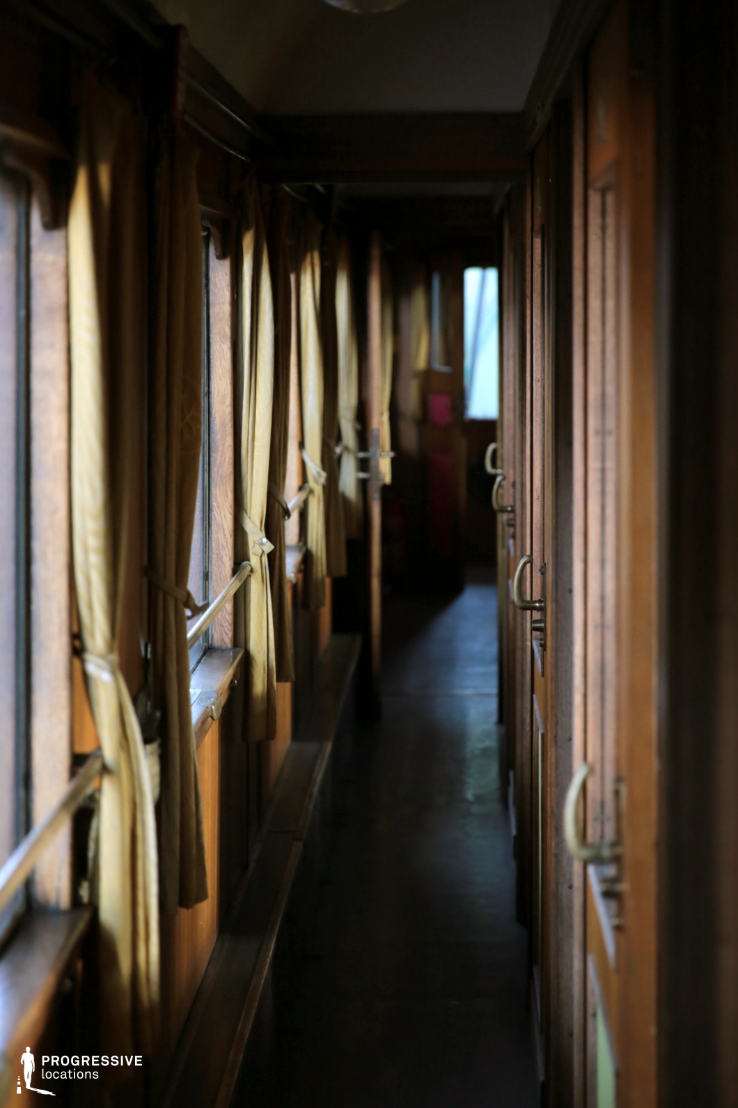 This dimly lit corridor inside a vintage railway carriage features a warm wooden interior with narrow passageways and individual cabin doors. The corridor is lined with tall windows, each adorned with golden fabric curtains, tied back to reveal glimpses of the outside. Metal handrails run along the walls, guiding passengers through the confined space. The rich wood paneling and brass door handles exude an old-world charm, ideal for historical or period-themed productions. The perspective of the corridor leads towards a softly illuminated doorway, enhancing the depth and nostalgic ambiance of the setting.