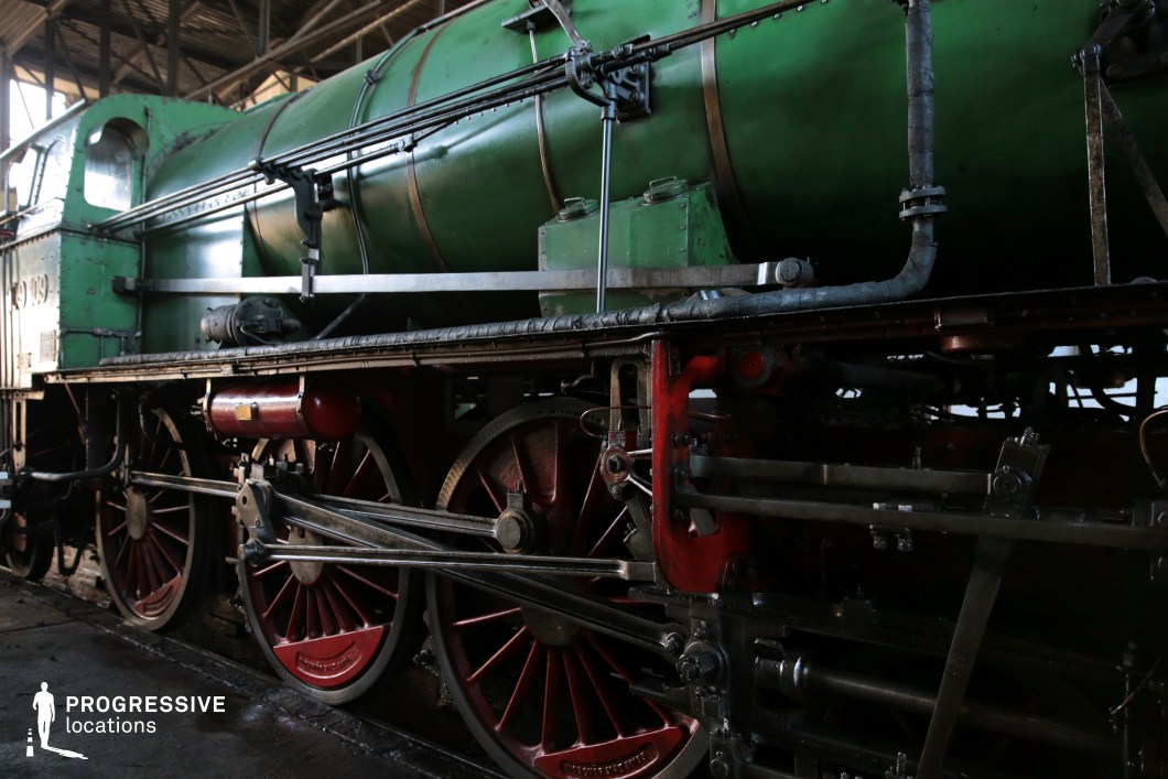 A close-up of a classic steam locomotive, featuring a green cylindrical boiler contrasted by red wheels and metal framework. The intricate mechanical components, including polished rods, pipes, and riveted panels, highlight the precision engineering of early railway transportation. The play of light and shadow across the worn yet well-maintained surface enhances the historical depth of the image. Ideal for period film productions, historical documentaries, and railway-themed artistic projects, this locomotive exudes both power and vintage charm.