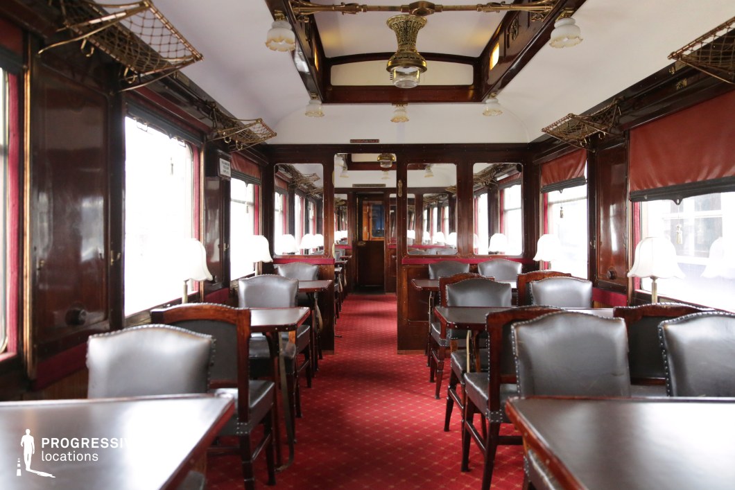 This historic train dining car exudes luxury with its dark polished wood paneling, deep red carpeting, and ornate brass light fixtures. The rows of black leather chairs and finely crafted wooden tables create a refined yet intimate setting, enhanced by soft-glowing table lamps that line the large windows. Overhead brass luggage racks and decorative ceiling elements further emphasize the carriage’s period charm. The symmetrical layout and reflective surfaces add depth and elegance, making this location ideal for period dramas, mystery thrillers, or classic travel-themed films requiring a setting of sophistication and nostalgia.
