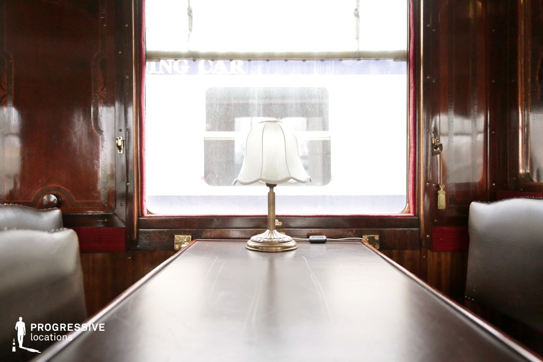 This close-up captures the refined elegance of a vintage train dining car, featuring a polished wooden table with a delicate, scalloped-edged table lamp as its focal point. The surrounding dark wood paneling, brass fixtures, and deep red accents contribute to the carriage's timeless luxury. Large windows allow soft natural light to filter in, subtly highlighting the craftsmanship of the space. The rich textures and symmetrical composition make this setting ideal for period dramas, mystery thrillers, or classic travel-themed films requiring an atmosphere of sophistication and nostalgia.