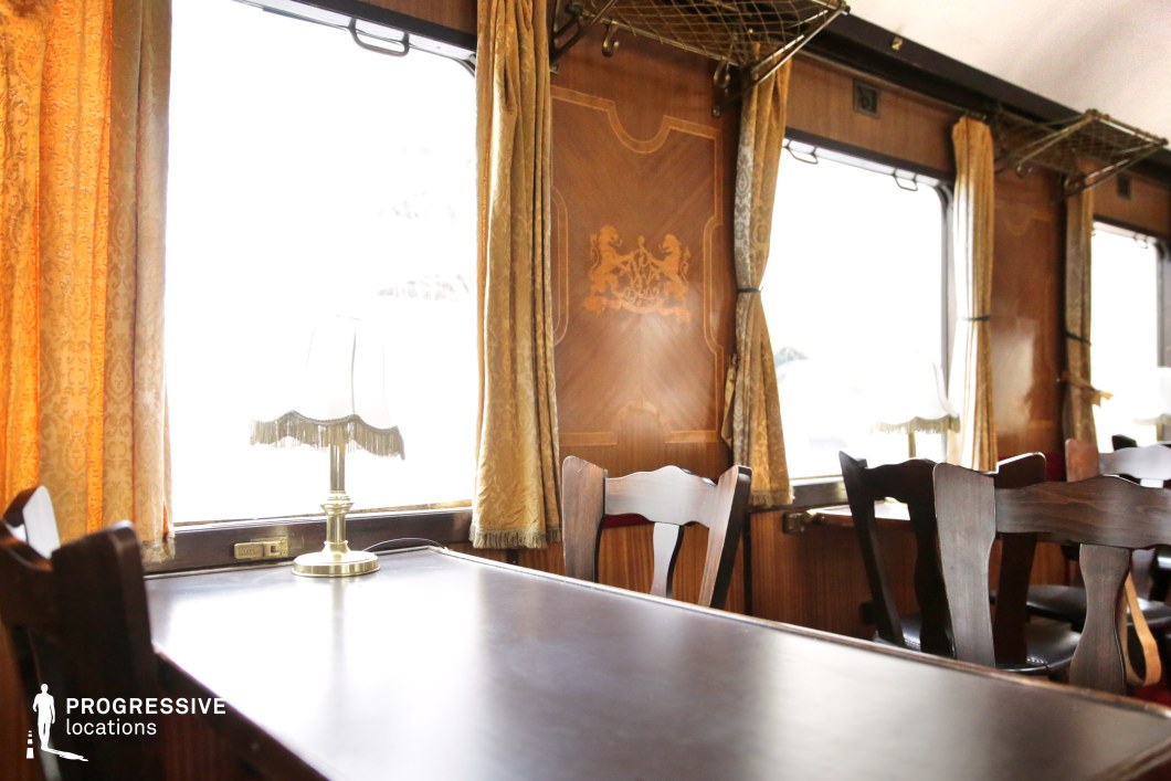 This close-up of a vintage train dining car highlights its elegant design, featuring dark polished wooden tables and chairs, rich golden drapes, and intricate wood paneling adorned with a decorative crest. A brass-framed table lamp with a fringed shade adds to the refined atmosphere, casting soft illumination over the setting. Overhead luggage racks and large windows contribute to the carriage’s classic luxury, creating an ambiance of nostalgia and sophistication. This setting is ideal for period dramas, historical thrillers, or travel-themed films that require a timeless and elegant railway environment.
