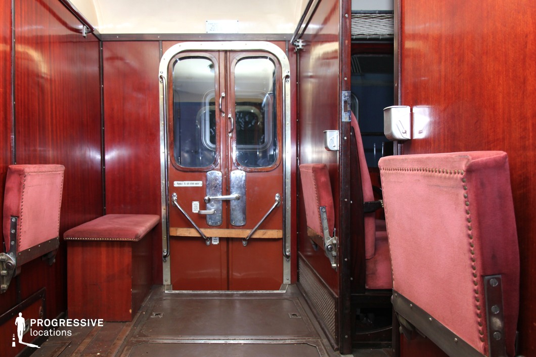 A classic train car interior with deep mahogany wood paneling and red velvet upholstered foldable seats. The central focus is the set of double doors with metal reinforcements and polished handles, leading to another section of the train. A small cushioned bench sits beside the entrance, adding to the vintage charm. The warm lighting reflects off the glossy surfaces, emphasizing the rich textures of the wood and fabric. Ideal for period film settings, evoking the atmosphere of mid-century railway travel with a sense of elegance and nostalgia.