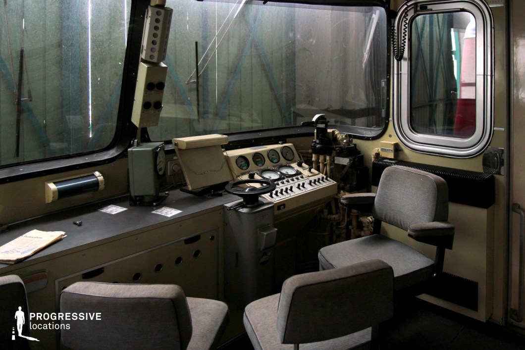 A functional and industrial-style train driving cab featuring a control panel with analog dials, switches, and levers. The cockpit is designed for operational efficiency, with a large wrap-around window providing a clear view of the surroundings. The muted beige and gray color scheme gives the space a utilitarian and retro aesthetic. The seating consists of cushioned gray chairs with armrests, positioned for both the operator and possible assistants. The overhead communication system and metal piping add to the mechanical character of the interior. This setting is ideal for historical, military, or industrial-themed productions requiring an authentic locomotive control room.