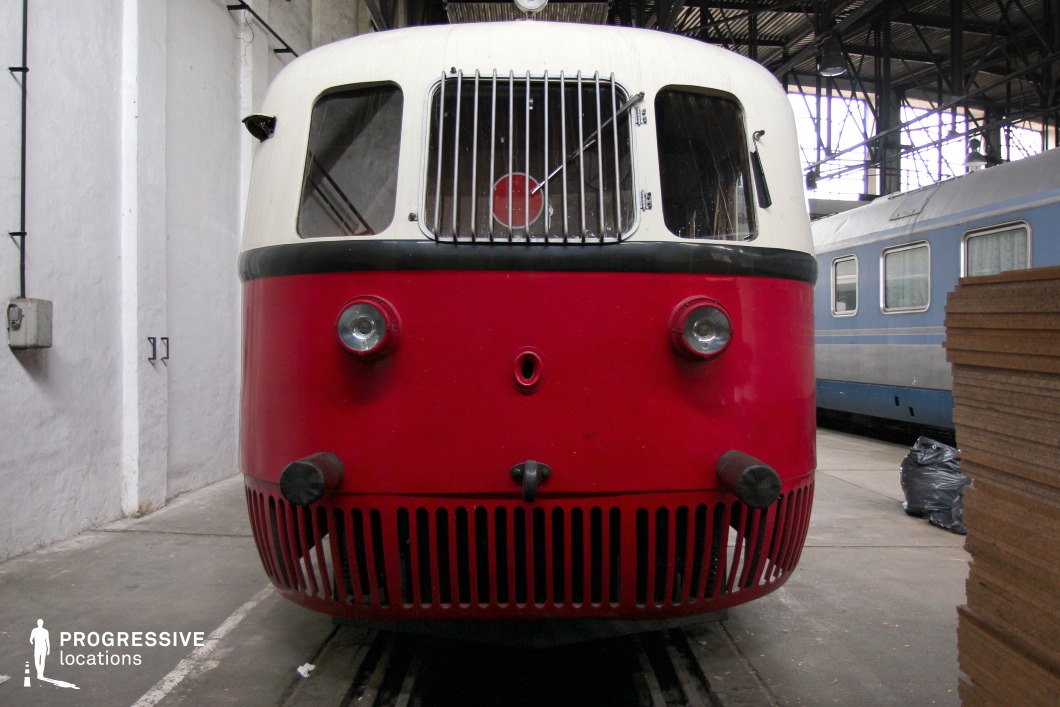 A vintage railcar with a distinctive red and white streamlined design, showcasing mid-20th-century engineering aesthetics. The rounded front features large windows for a panoramic driver's view, protected by a metal grille. Dual headlights and black cylindrical buffers emphasize its functional yet charming appearance. The lower section is adorned with vertical air vents, adding to its retro-futuristic look. The train is housed inside an industrial depot with exposed steel beams and concrete walls, enhancing the nostalgic ambiance. This setting is ideal for historical or period-specific film projects, evoking mid-century railway travel with a unique design.