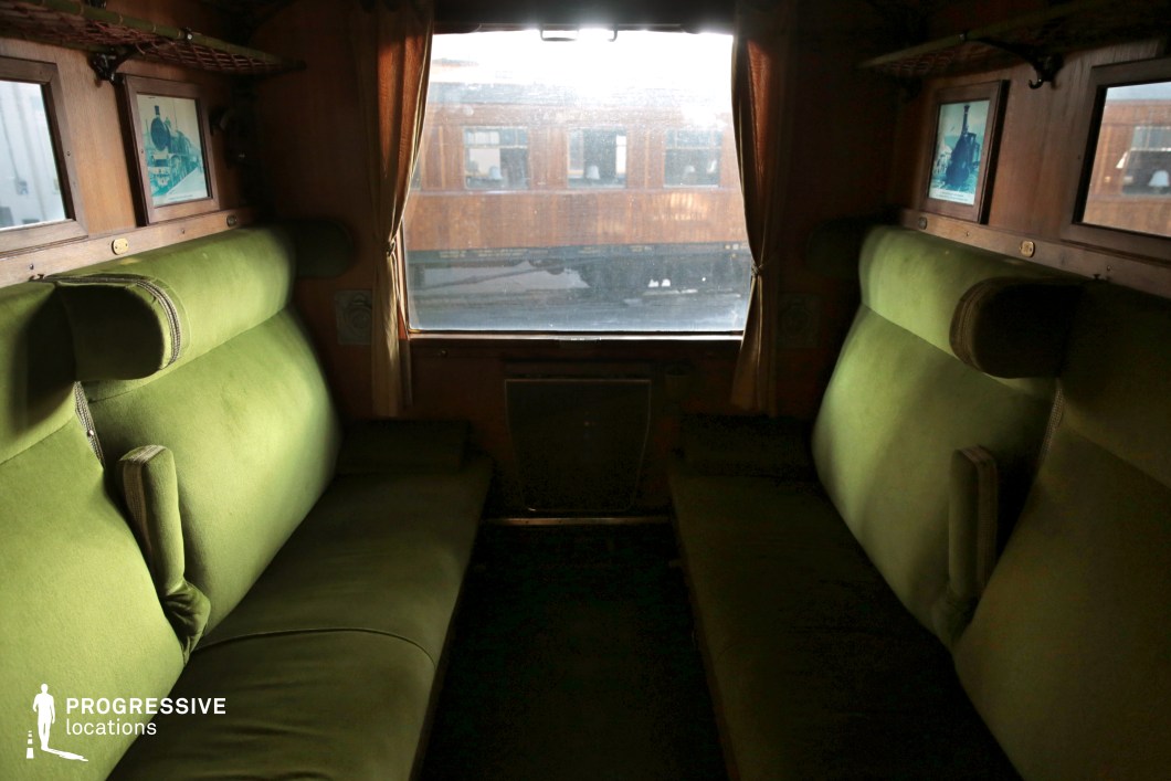 A classic train compartment featuring two opposing green upholstered bench seats with plush cushioning and integrated armrests. The wooden-paneled walls are adorned with framed vintage locomotive prints, enhancing the historic ambiance. Above the seats, metal luggage racks provide storage space. A large central window, framed by delicate golden curtains, allows natural light to illuminate the cabin, offering a glimpse of another vintage wooden train car outside. The dark flooring contrasts with the warm-toned wood and green upholstery, creating an elegant and cozy setting reminiscent of early 20th-century railway travel.