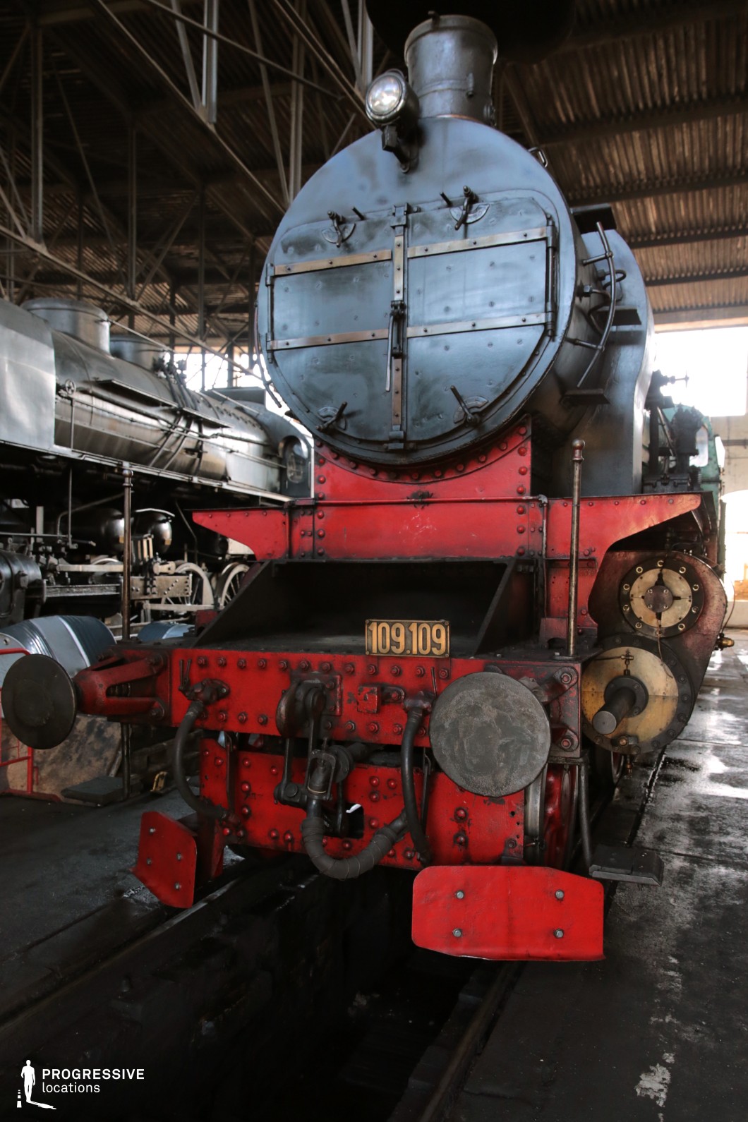 A striking front view of a historic steam locomotive, featuring a bold black boiler and red undercarriage. The detailed metalwork, rivets, and industrial components reflect the robust engineering of early 20th-century railway design. Positioned in an indoor depot with other locomotives visible in the background, the image highlights the locomotive's utilitarian strength and mechanical precision. The worn yet well-preserved exterior adds to the authenticity of the setting, making it ideal for period film productions, industrial photography, and transportation history projects.