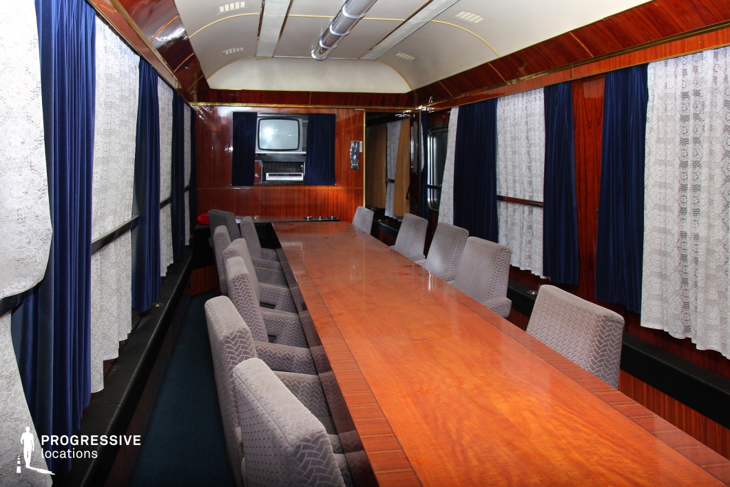 A refined and formal meeting space inside an armored train carriage, featuring a long polished wooden table surrounded by upholstered chairs in muted gray tones. The rich mahogany-paneled walls enhance the elegant and retro aesthetic, complemented by navy blue velvet curtains paired with delicate white lace drapery. Overhead, a curved ceiling with metal ventilation fixtures reinforces the functional yet sophisticated design. A vintage television and air conditioning unit are mounted at the far end, hinting at mid-to-late 20th-century origins. This unique setting is ideal for historical, political, or military-themed productions requiring a high-level discussion atmosphere within a moving command center.