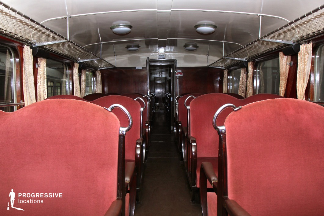 A classic mid-20th-century passenger train car with a structured, symmetrical seating arrangement featuring plush red upholstered chairs with curved metal armrests. The dark wooden paneling and chrome accents enhance the period aesthetic, while the cream-colored ceiling with round, evenly spaced lights provides a soft illumination. Overhead luggage racks with metal mesh detailing line both sides, and delicate beige curtains frame the large windows, allowing natural light to filter in. This train interior exudes a retro, cinematic feel, making it ideal for period films or nostalgic travel sequences.
