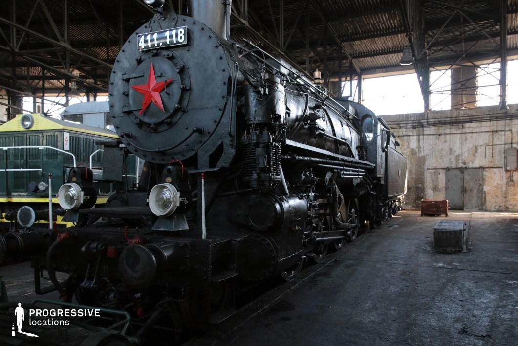 A powerful black steam locomotive with a striking red star emblem on its front is parked inside an industrial railway depot. The locomotive's robust metal construction, intricate piping, and large headlights reflect the engineering of mid-20th-century railway design. The worn concrete walls, exposed pipes, and metal framework of the depot create a gritty, historical atmosphere. A green diesel locomotive is partially visible in the background, adding contrast to the setting. This location is ideal for historical and war-era productions, evoking themes of industrial power, Soviet history, and Cold War aesthetics.