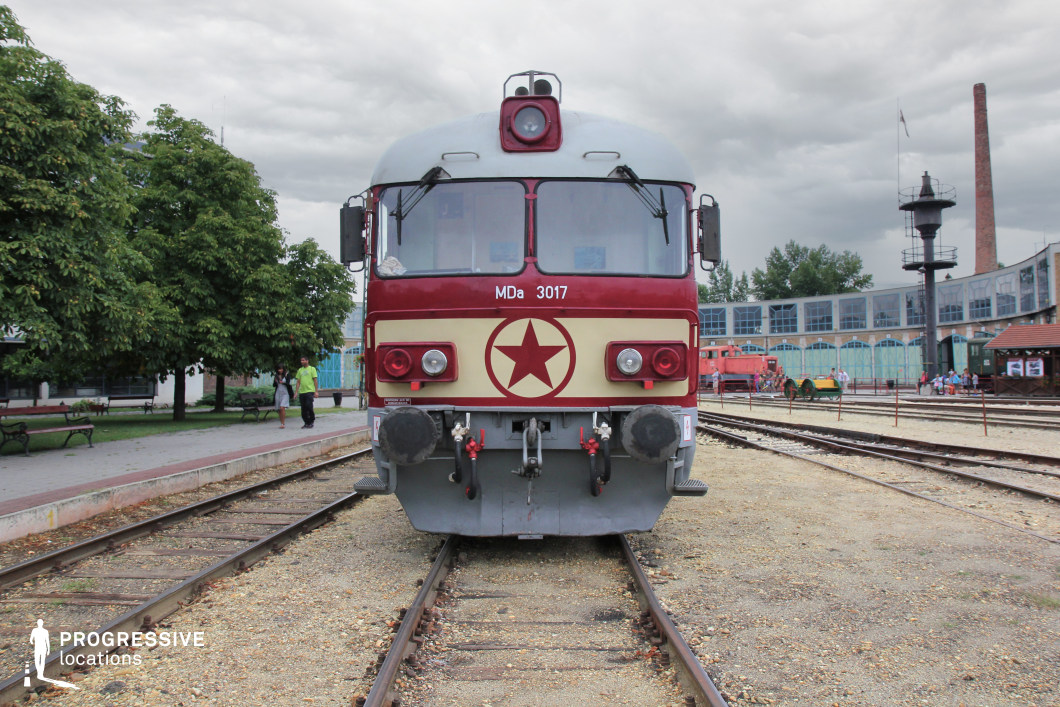 A frontal view of a classic diesel locomotive, painted in a striking red and cream color scheme, featuring a prominent red star emblem. The design reflects mid-to-late 20th-century socialist-era railway aesthetics. The locomotive is stationed in an open-air railway museum, surrounded by historical buildings, industrial-era infrastructure, and various vintage train cars in the background. The setting provides a nostalgic yet imposing atmosphere, ideal for historical reenactments, Cold War-era film productions, or transportation-themed visual projects.