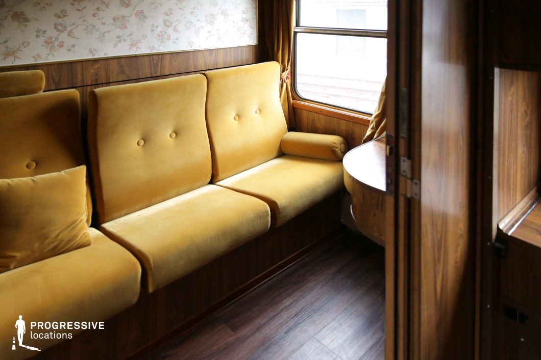This inviting train cabin features plush mustard-yellow seating with a retro button-tufted design, paired with a warm wooden interior. The floral-patterned wallpaper adds a nostalgic charm, complementing the classic train aesthetic. A built-in table by the window enhances functionality, while the large window allows natural light to illuminate the space. The overall ambiance suggests comfort and refinement, making this location ideal for period dramas, intimate travel stories, or mystery thrillers requiring a stylish and historically rich train setting.