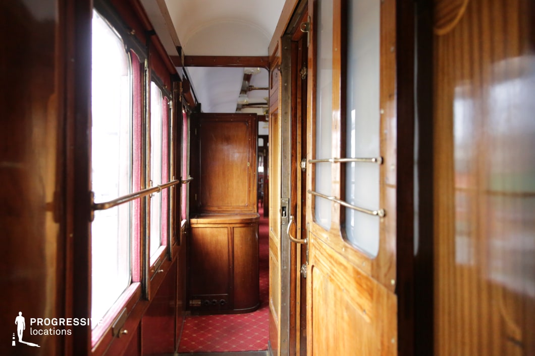 This classic train car corridor is lined with polished wooden paneling, brass railings, and frosted glass cabin doors, evoking the refined elegance of early 20th-century rail travel. The deep red carpeting contrasts with the warm wood tones, while natural light filters through the large windows framed by rich red curtains. The slightly curved ceiling and narrow passage enhance the carriage's intimate and immersive atmosphere. This location is ideal for period dramas, suspenseful thrillers, or mystery films requiring a nostalgic and historically detailed train setting.