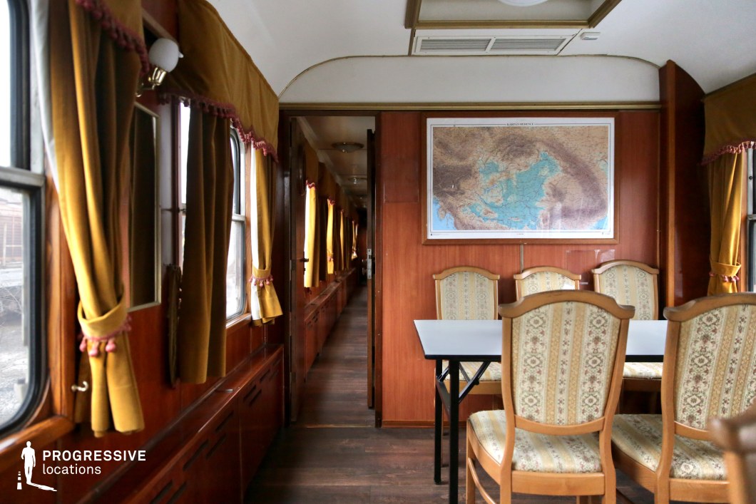 This vintage train salon car features a refined wooden-paneled interior with an intimate dining or meeting space. The soft golden drapes with decorative tassels frame large windows, enhancing the warm and nostalgic ambiance. The seating area consists of elegant upholstered chairs with intricate patterns, arranged around a simple table. A large framed map adorns the back wall, adding a historical touch. The narrow corridor leading further into the carriage creates depth and perspective, reinforcing the classic rail travel aesthetic. This setting is ideal for period dramas, political thrillers, or character-driven narratives that require a refined yet nostalgic train interior.
