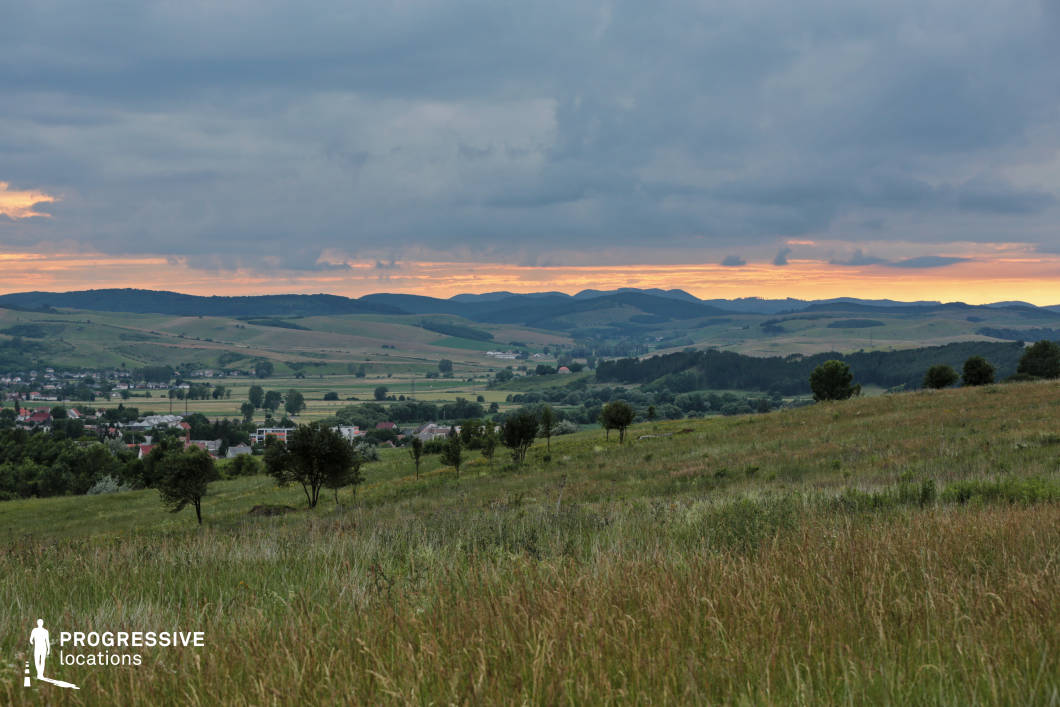 A scenic view of gently sloping meadows transitioning into a patchwork of farmland and a small village nestled in the valley below. The distant rolling hills provide a striking backdrop, their silhouettes adding depth to the landscape. Scattered trees dot the foreground, enhancing the rural charm and natural texture of the grassy field. The blend of open space, agricultural fields, and a small settlement highlights the harmonious coexistence of nature and human habitation. This location is ideal for portraying tranquil rural life, wide open spaces, and the beauty of undisturbed natural surroundings.