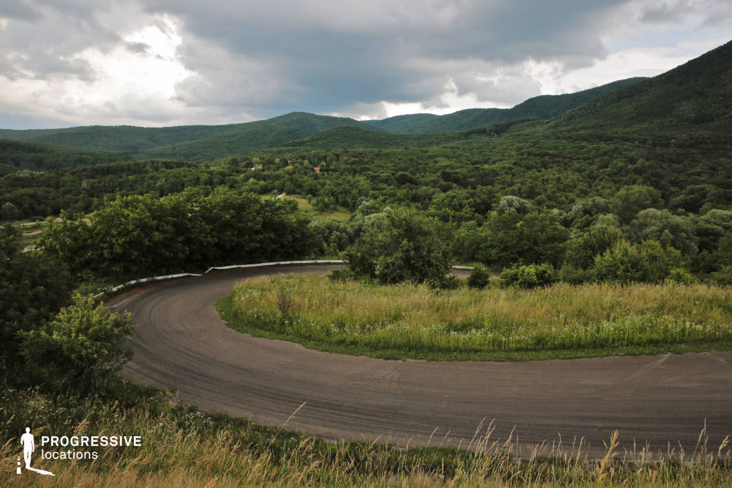 The image showcases a striking 160-degree bend in a narrow serpentine road, surrounded by rolling, forested hills that stretch across the horizon. The sharp curve, bordered by wild grasses and low shrubs, commands attention as it snakes dramatically through the vibrant green landscape. The dense foliage, with varying shades of green, forms a rich, natural backdrop that contrasts with the dark, weathered asphalt of the road. This steep, hairpin turn is ideal for cinematic scenes emphasizing challenging routes, remote wilderness, or the rugged beauty of untamed nature.