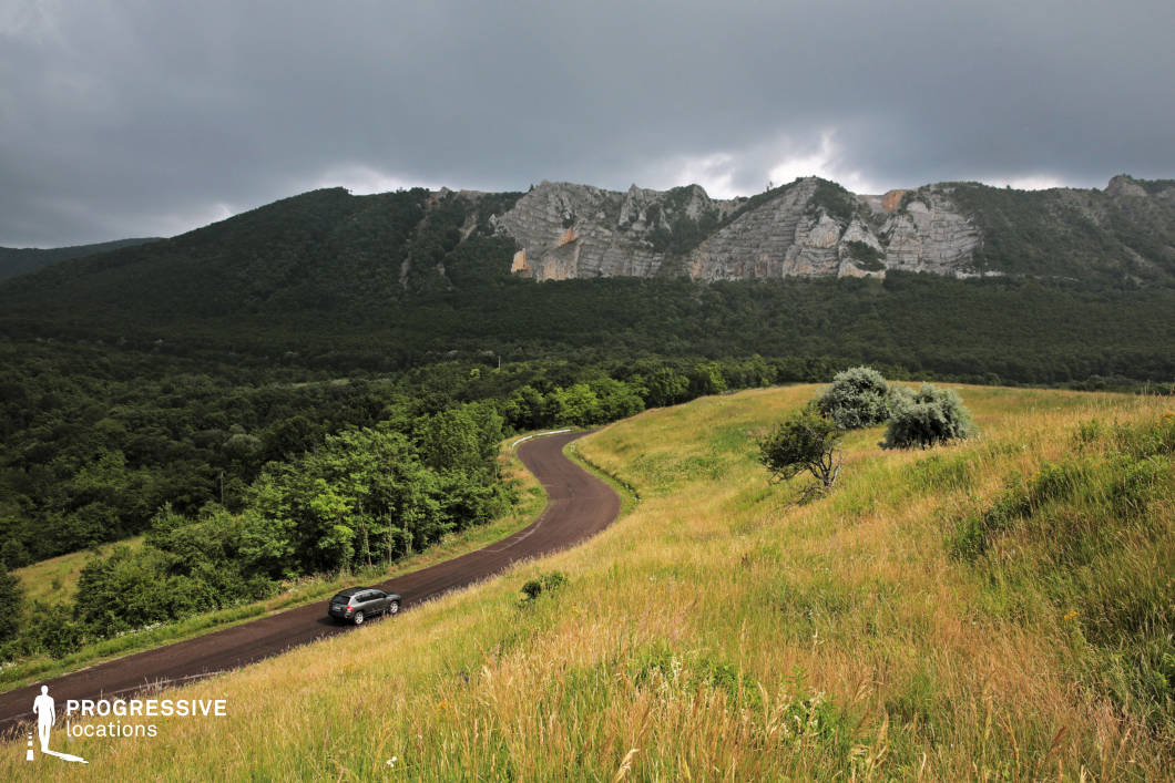 A striking view of a winding road traversing an open, grassy hillside with towering rocky cliffs in the background. The cliffs' rugged formations rise above the lush greenery, providing a dramatic natural contrast. The road's smooth curve and the lone car highlight the remote, untamed beauty of the scene. The landscape’s vibrant green tones, combined with the dramatic cliffs, make this an ideal setting for adventurous or cinematic visuals emphasizing nature's grandeur and raw beauty.