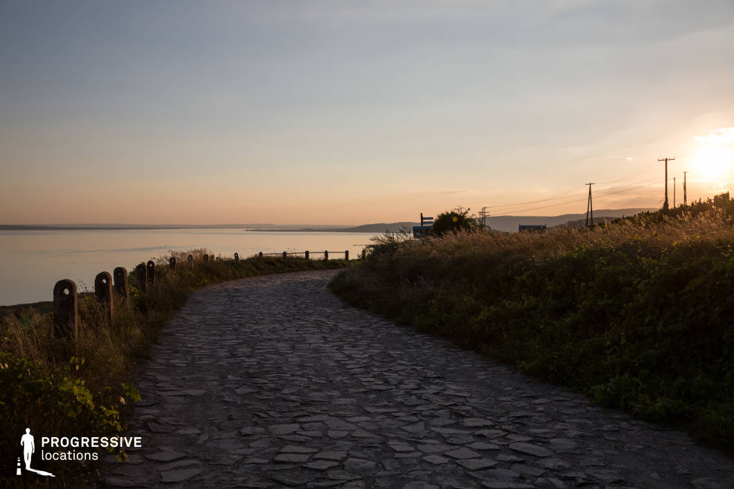 This serene image depicts a cobblestone road curving gently along the edge of a tranquil lake, bathed in the warm hues of sunset. The road is bordered by grassy embankments and rustic wooden posts, enhancing its natural charm. The calm water and distant hills blend harmoniously with the soft golden light, creating a peaceful and nostalgic atmosphere. Ideal for scenes emphasizing reflection, nature, or quiet rural beauty.