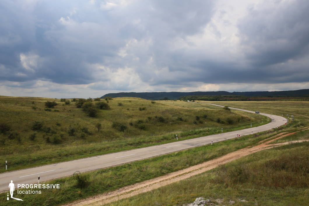 This panoramic view captures a winding rural road cutting through expansive grassy fields, with gentle hills rolling into the distance. The light and shadow of the cloudy sky enhance the mood of the landscape, balancing drama and stillness. The road's gentle curves and the untouched countryside evoke a sense of journey and exploration. An ideal setting for showcasing freedom, adventure, or the beauty of wide-open spaces.