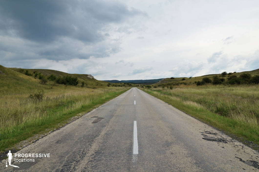 This image captures a straight, two-lane country road extending into the horizon, framed by expansive grassy hills on both sides. The road's worn asphalt texture, with visible cracks and faded markings, adds a rustic and authentic feel to the scene. The surrounding greenery consists of tall grass and scattered shrubs, creating a natural and unmanicured landscape. Gentle rolling hills provide subtle elevation changes, and the muted color palette of greens and grays establishes a calm and desolate atmosphere. The perspective draws the eye directly down the road, emphasizing a sense of depth and openness. This location is ideal for filming scenes with themes of solitude, travel, or transition.