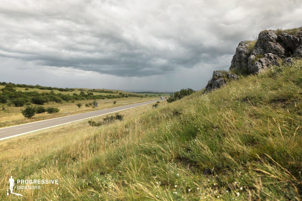 This image features a gently sloping hillside adorned with wild grasses and dotted with rugged rock formations. In the background, a straight asphalt road cuts through the open landscape, surrounded by vast fields and sparse vegetation. The muted tones of the rocks and grass contrast with the expansive cloudy sky, lending a dramatic yet serene atmosphere. The open view and natural elements make this an ideal location for wide-angle shots or establishing scenes showcasing nature’s raw beauty.
