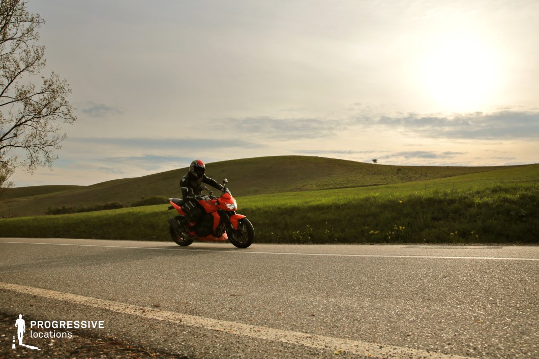 This image showcases a smooth, open country road meandering through a landscape of gently rolling green hills. The road’s well-maintained surface is bordered by lush grass and wildflowers, which enhance the rural charm. The hills stretch across the horizon, creating a sense of depth and serenity. The soft light and expansive sky complete the tranquil atmosphere, making this location ideal for emphasizing the beauty of open spaces and quiet journeys.