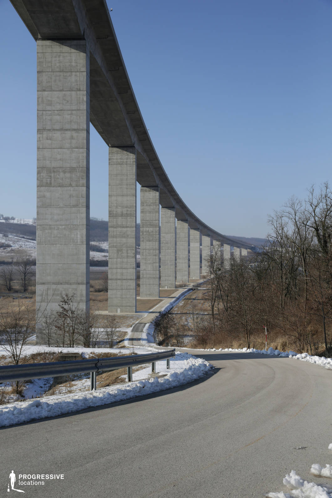 This location centers on a winding rural road running parallel to a towering viaduct, which serves as a striking visual backdrop. The road, bordered by metallic guardrails and patches of snow, provides a functional and accessible space for various human activities or narratives to unfold, such as a journey, an encounter, or a pursuit. Its gentle curves create a natural flow and rhythm, adding visual dynamism to the composition. The viaduct, with its massive concrete supports and sweeping, elevated structure, amplifies the setting's dramatic appeal. Its monumental presence introduces a contrasting verticality and industrial texture to the otherwise tranquil rural landscape, making it a unique and visually compelling element that enriches the scene. This interplay between the grounded road and the imposing viaduct above makes the location particularly versatile for film and photography, offering opportunities for striking contrasts and dramatic storytelling.
