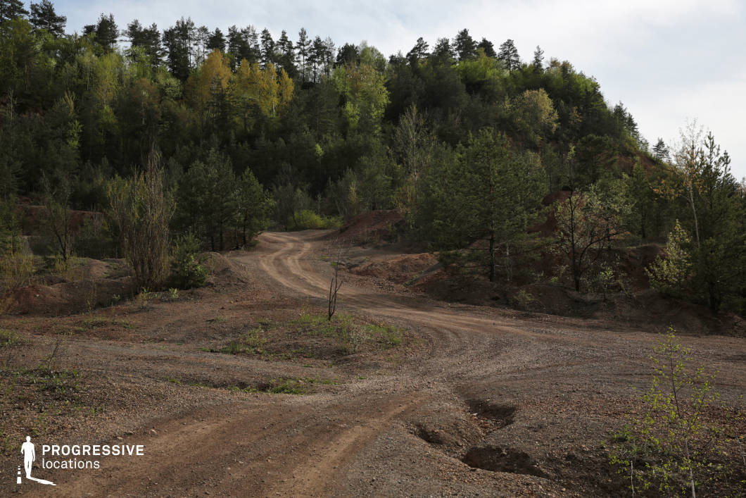 This rugged gravel road winds through a forested landscape, offering a raw and untamed setting ideal for scenes that emphasize exploration, isolation, or adventure. The uneven surface and loose gravel suggest a location that sees little maintenance, enhancing its untamed and naturalistic character. Surrounded by dense greenery and a mix of young and mature trees, the road meanders through the clearing, creating a sense of depth and direction. The subtle elevation changes in the terrain and the reddish-brown soil add texture and visual interest. This location provides a versatile backdrop for off-road sequences, outdoor activities, or moments of quiet reflection in a secluded natural environment.