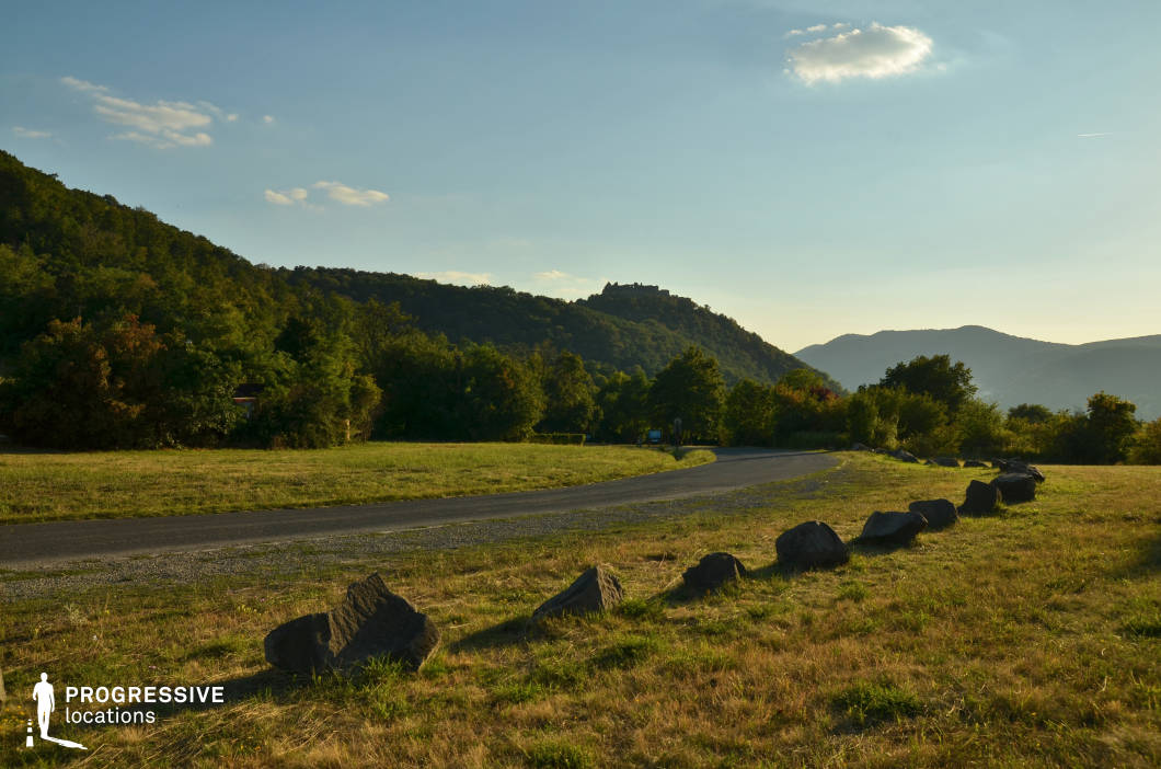 This location features a gently curving road set amidst a picturesque hillside landscape, surrounded by lush greenery and distant forested slopes. Large rocks line the edge of the road, creating a natural boundary and adding texture and detail to the scene. The grassy field in the foreground transitions smoothly into a wooded area, with rolling hills and a distant hilltop castle visible on the horizon, providing a dramatic focal point and a touch of historical intrigue. The warm, golden light bathes the landscape, enhancing the sense of tranquility and openness. This location is ideal for capturing moments of calm exploration or scenic journeys, offering ample space for action along the road and visual depth created by the layered elements of hills, trees, and distant structures.