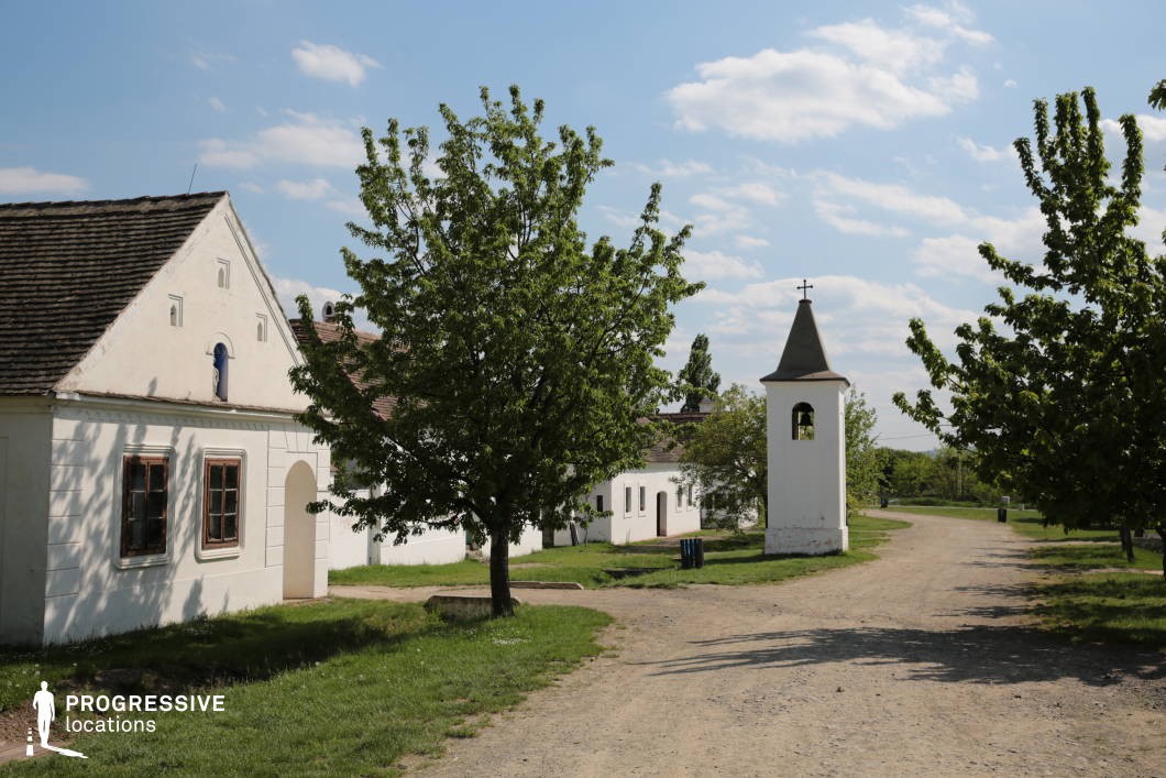 This village setting showcases a picturesque dirt road flanked by traditional whitewashed buildings with tiled roofs, capturing the essence of rural heritage. The buildings exhibit a historic, modest architectural style, featuring small wooden-framed windows and arched doorways, adding to their old-world charm. One structure has a small religious statue embedded in its gable, hinting at cultural or historical significance. A small bell tower with a pointed roof and a metal cross stands prominently along the roadside, reinforcing the village’s serene and historical character. The unpaved road meanders gently, bordered by lush greenery and mature trees, which cast natural shadows and enhance the depth of the scene. This location presents an authentic countryside atmosphere, ideal for film productions or photography projects requiring a peaceful, historically resonant rural environment. The open road, framed by scattered trees and rustic buildings, allows for dynamic compositions, making it suitable for period pieces, documentaries, or scenic landscape captures.