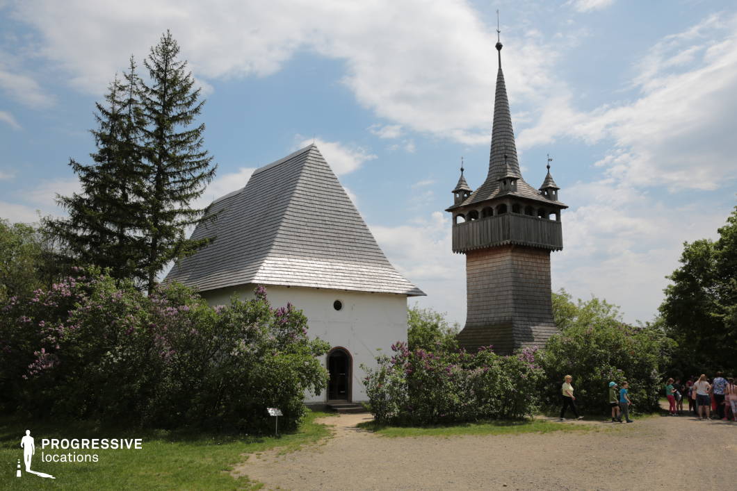 This setting features a remarkable wooden church tower with an intricately designed spire, standing adjacent to a simple white chapel with a steeply pitched wooden-shingled roof. The tower, built in a traditional Eastern European style, exhibits fine craftsmanship with its open belfry and pointed, ornate turrets, evoking historical and cultural significance. Surrounding the structures is lush greenery, including tall evergreen trees and vibrant lilac bushes, adding a natural frame to the composition. The unpaved pathway leading to the entrance enhances the rural and historical character of the location, while the soft contrast between the dark wooden tower and the bright white chapel creates a visually appealing juxtaposition. This location is ideal for film productions or photography projects that seek to capture historical, religious, or folk-inspired settings. The architectural details and natural elements provide a serene and picturesque backdrop, making it suitable for period dramas, cultural documentaries, or rural storytelling.