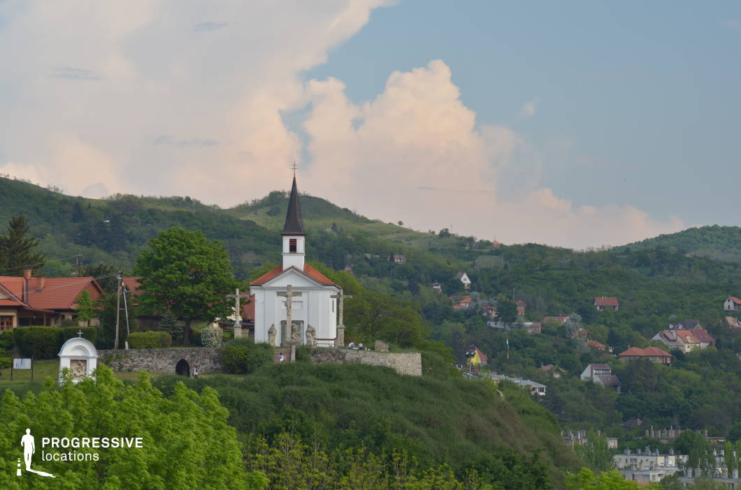 Perched atop a scenic hill, this picturesque chapel features a traditional white facade, a sharply pointed steeple with a red-tiled roof, and a small bell tower that adds to its historical charm. A set of religious statues and stone crosses in front of the church create a serene and spiritual ambiance, further emphasizing the sacred nature of the site. The low stone wall surrounding the area enhances the sense of elevation and separation from the landscape below. The background consists of rolling green hills dotted with houses and traditional countryside dwellings, adding depth and a strong rural character to the composition. The elevated position of the chapel provides a commanding view of the surrounding landscape, making it an excellent location for scenic photography, historical documentaries, or film productions that require a tranquil yet dramatic setting. The combination of religious architecture, natural elevation, and lush greenery makes this an ideal location for projects focusing on heritage, spirituality, or breathtaking panoramic countryside visuals.