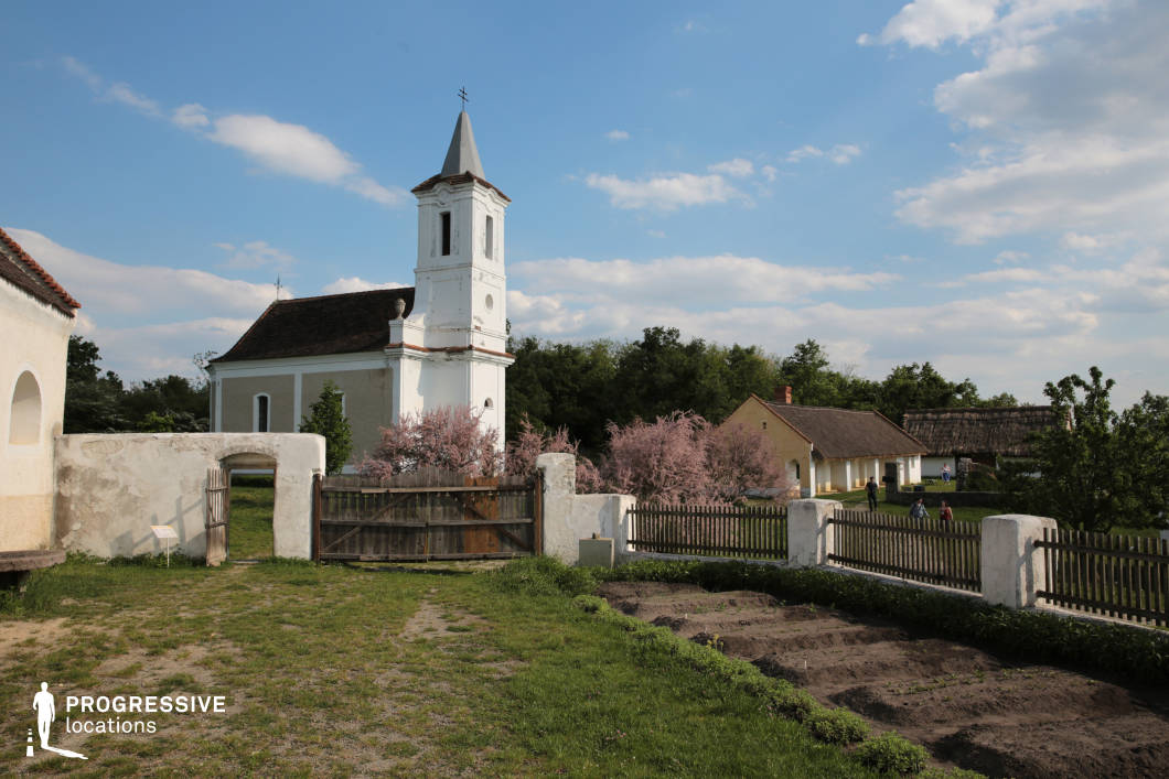 This serene rural scene features a modest, historic church with a white bell tower and a pointed roof, enclosed by a rustic wooden gate and a low plastered wall. The church's classic architecture, with a pitched roof covered in weathered tiles and subtle decorative elements on its facade, evokes a timeless countryside aesthetic. In the foreground, a small cultivated garden with freshly tilled soil adds an element of traditional village life. The fence surrounding the area is a mix of wooden pickets and stone pillars, reinforcing the rural authenticity of the location. Pink blossoming trees near the church soften the composition, adding vibrancy and a seasonal touch. Beyond the church, traditional thatched-roof and plastered village houses blend harmoniously into the landscape, surrounded by rolling greenery and mature trees. This setting offers a visually rich and historically resonant atmosphere, making it ideal for film and photography projects focused on heritage, countryside life, or European rural culture.