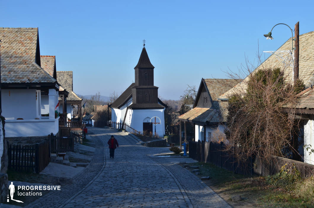This picturesque village street, paved with aged cobblestones, winds gently through a collection of traditional whitewashed houses with steeply pitched, weathered-tiled roofs. The structures exhibit a folk architectural style, featuring wooden balconies, dark timber framing, and neatly arranged facades that reflect a strong historical character. Dark wooden fences and natural elements such as leafless vines add to the rustic charm of the setting. A distinctive feature of the scene is the wooden church with a dark timber-clad bell tower, standing at the focal point of the street. The contrast between its white base and dark upper structure makes it visually striking. The surrounding landscape gently slopes downward, providing a sense of depth and perspective. This location is ideal for film productions or photography projects seeking a preserved historical atmosphere, making it suitable for period pieces, cultural documentaries, or heritage tourism visuals. The combination of traditional architecture, natural materials, and a gently curving street enhances the authenticity of the rural European village aesthetic.