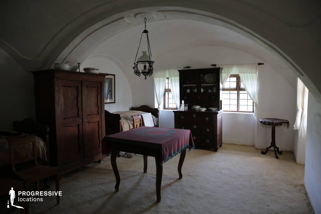 This traditional village home interior features a white vaulted ceiling with elegant curved arches, creating a sense of spaciousness while maintaining a cozy, enclosed atmosphere. The natural light filtering through small-paned wooden windows, framed by delicate white curtains, enhances the soft and timeless ambiance of the space. The room is furnished with antique wooden furniture, including a dark-finished armoire, a dining table draped with a patterned textile, and a classic wooden sideboard displaying neatly arranged porcelain dishes. Two wooden-framed beds with carved headboards and traditional embroidered blankets reinforce the historic rural aesthetic. The hanging metal lantern adds an authentic period touch, contributing to the room’s warm and subdued lighting. The unpolished, earth-toned flooring complements the rustic simplicity of the space, emphasizing its historic and functional nature. This location is ideal for period film settings, historical documentaries, or photography projects that require an authentic representation of traditional rural life. The interplay of aged materials, simple elegance, and soft natural light makes this a compelling interior for heritage and cultural storytelling.